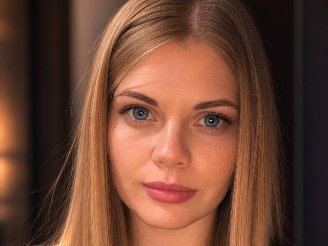 Pannia has straight, blonde hair, blue eyes, and pink lips. Her face is centered with a neutral expression, showing subtle makeup and a smooth complexion.