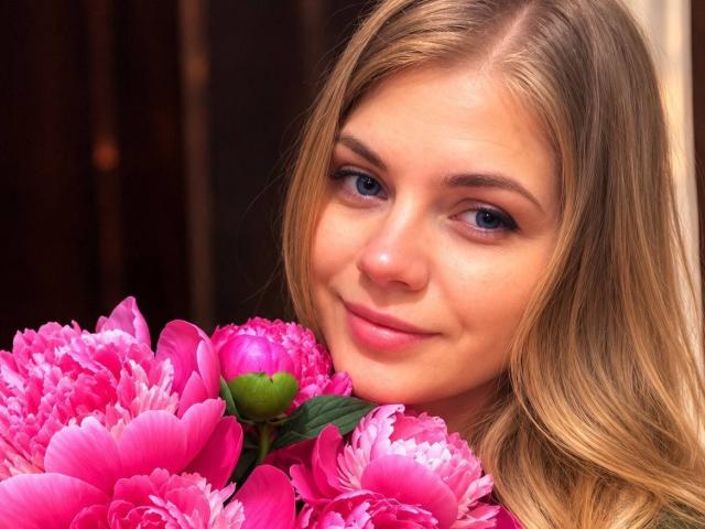 Pannia with blonde hair and blue eyes, smiling softly, holding pink peony flowers, her nose and lips slightly upturned, a green peony bud visible in the bouquet.