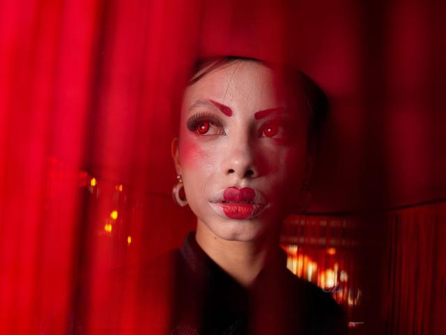 Kazumii has red eyes, bold red eyebrows, and red lipstick, with red curtains partially obscuring her face. She wears a black top and has small hoop earrings.