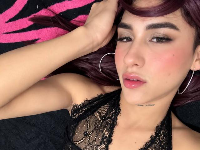 SinfulDuo with purple hair, hoop earrings, and pink lipstick, wearing a black lace halter top, lying down, hand in hair, small tattoo under chin, slightly flushed, sultry expression.