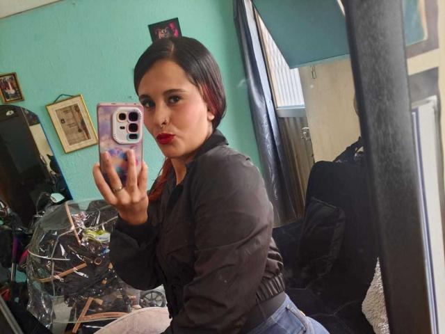ChandyLove with short black hair, red lipstick, black jacket, and blue jeans, taking a mirror selfie, showing her ass, holding a phone with a colorful case.