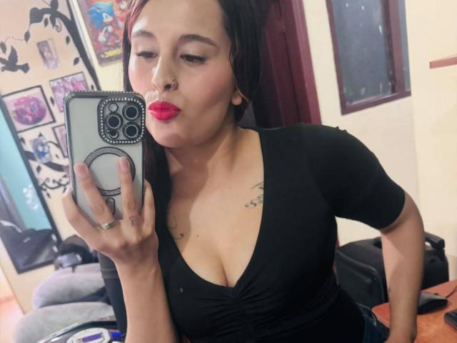 ChandyLove with dark hair, red lipstick, nose ring, black top showing her boobs, taking a selfie in a mirror, holding a phone with a ring, in a room.