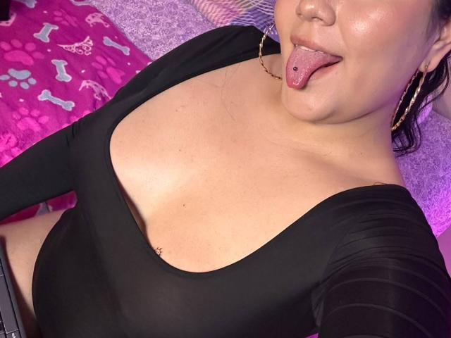 ArieWilden with tongue out, black top, showing big tits, pierced tongue, gold hoop earrings, and tan skin, lying on pink bone-patterned bed.