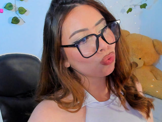 JennyLawry with pouty lips, wearing black glasses, white halter top, and long brown hair, sitting in a black chair, next to a large tan teddy bear.