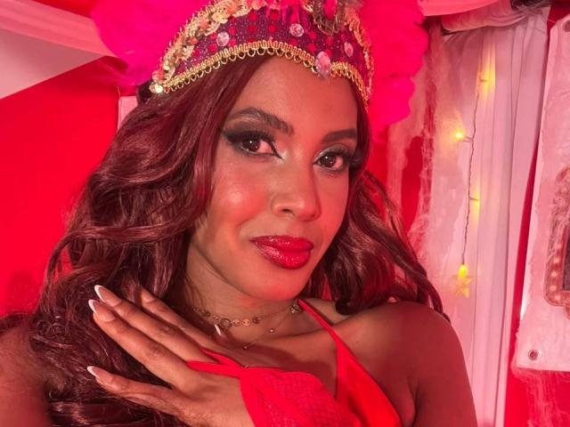 CamilaWest con rossetto rosso, capelli ricci castano scuro e una corona dorata e rosa, indossa un abito rosso, con una mano sul petto, sorridendo con un trucco marcato..