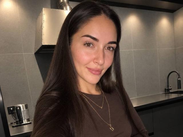 AquaLady, con cabello castaño largo, ojos marrones y una nariz pequeña, vestida con una camisa marrón y un collar dorado, sonriendo en una cocina moderna. Sus senos no son visibles..