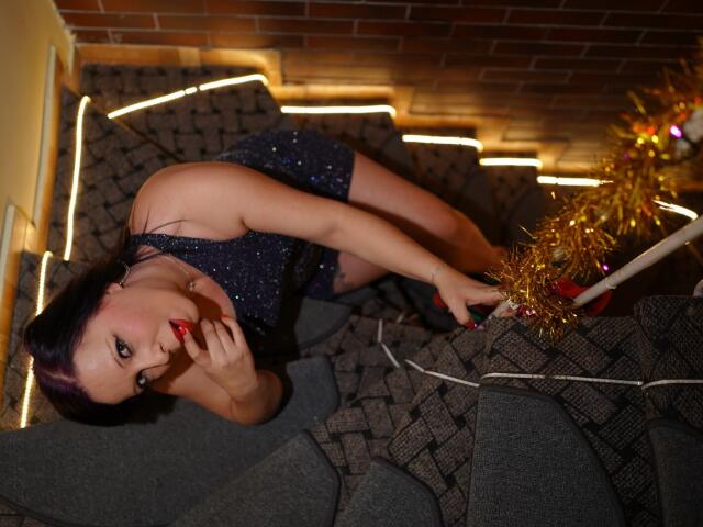 LauraSin with purple hair, red lipstick, and sparkly blue dress, lying on patterned gray couch, looking up, holding gold tinsel and white ribbon, fingers near her glossy lips.