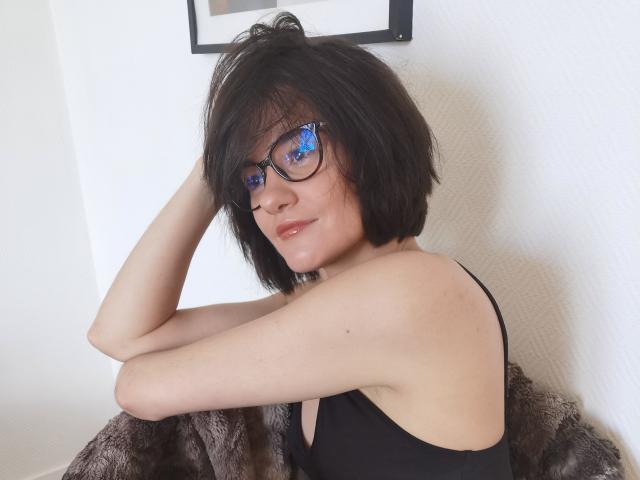 FrancaisePhoenix with short black hair, blue reflection glasses, black tank top, and arms raised, showing armpits, sitting on a furry brown blanket.