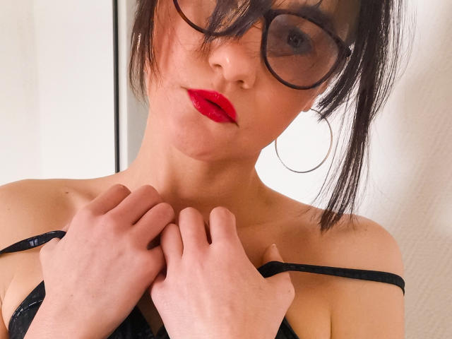 FrancaisePhoenix with red lipstick, black glasses, and black strap lingerie, holding straps, showing her tits, white wall background, and large hoop earrings, looking into camera.