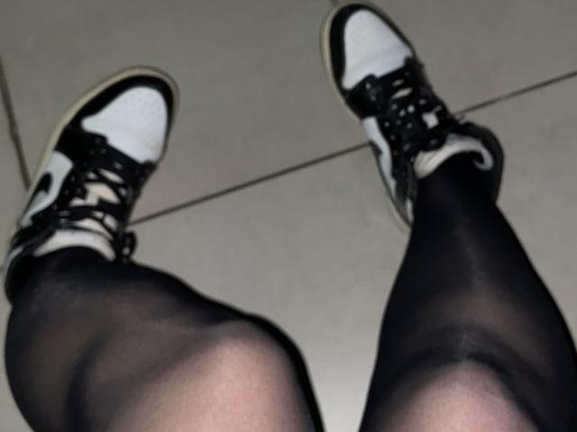 Le gambe di Tinelia indossano collant neri trasparenti e scarpe da ginnastica bianche e nere, con i lacci allacciati e le punte dei piedi leggermente rivolte verso l'esterno. Le scarpe da ginnastica hanno suole nere e dettagli bianchi..