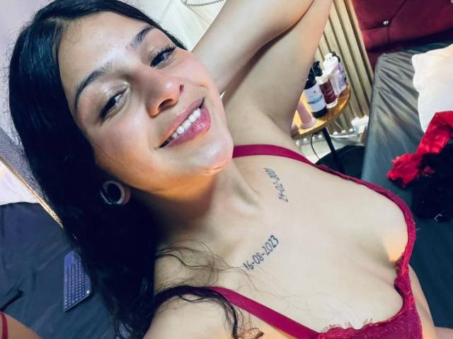 LonxEva with long black hair, smiling, wearing a red lace bra, showing small tattoos above her tits, and large gauged earrings, lying on a bed, arm raised.