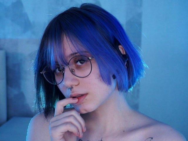 YasukoCella mit blau gefärbten Haaren, Brille, Nasenring und schwarzem Labret-Piercing, beißend auf ihren Finger, verführerisch, oben ohne, mit freiliegenden Schlüsselbeinen. Blauer Schein, kurzer Bob, süß, sexy..