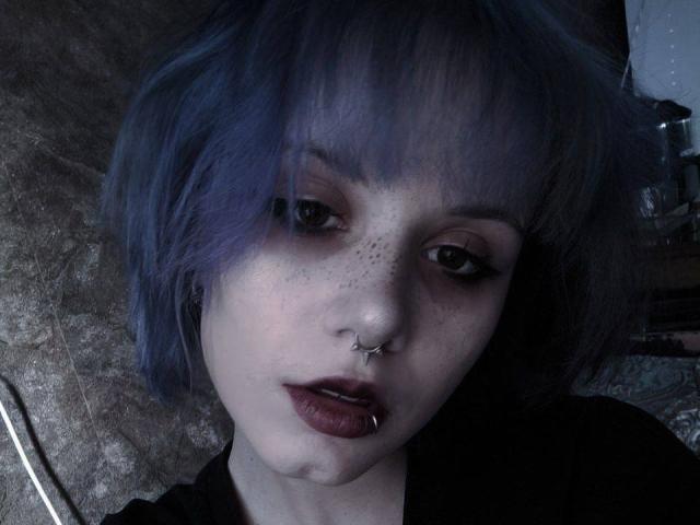 YasukoCella tem o cabelo tingido de azul, um piercing no nariz e um piercing no lábio. Ela é pálida, com sardas, delineador escuro e batom roxo. Sua boca está ligeiramente aberta, olhando para cima..