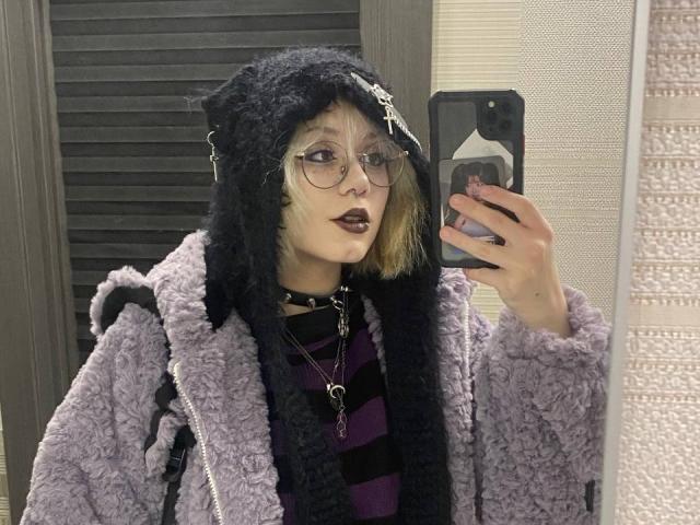 YasukoCella mit schwarzem Wollhut, Brille, lila Lippenstift, lila gestreiftem Hemd, schwarzem Choker, lila, flauschiger Jacke, hält ein Handy und macht ein Selfie im Spiegel, blonde Haare, schwarzer Rucksack..