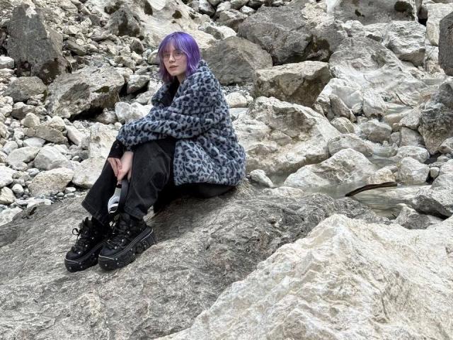 YasukoCella with purple hair, gray leopard print coat, black pants, and black chunky shoes, sitting on rocky terrain, looking at camera with subtle smile. Rock textures in foreground.