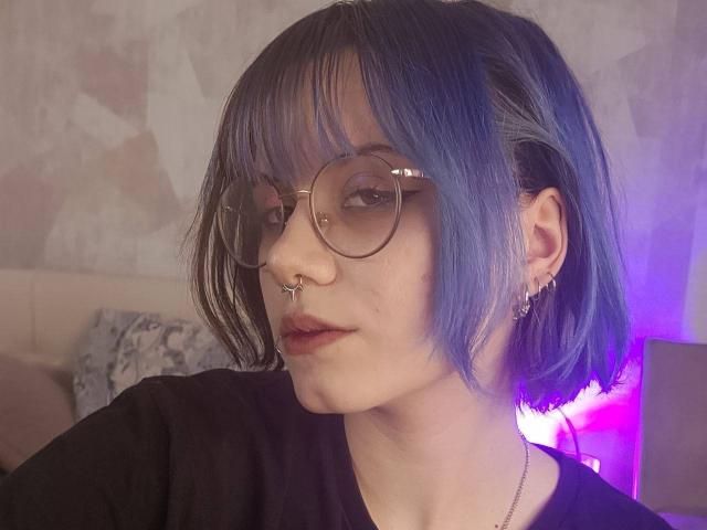 Yasuko, con caschetto biondo, occhiali rotondi, anello al naso, piercing al setto, piccoli orecchini, maglietta nera, labbra leggermente serrate, luce viola tenue, capelli corti e disordinati, pelle pallida, che guarda di lato..