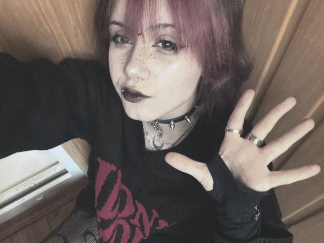 YasukoCella with pink-dyed hair, black lipstick, nose ring, and septum piercing, wearing a black shirt with red design, spiked collar, and ring, hand up, looking at camera.