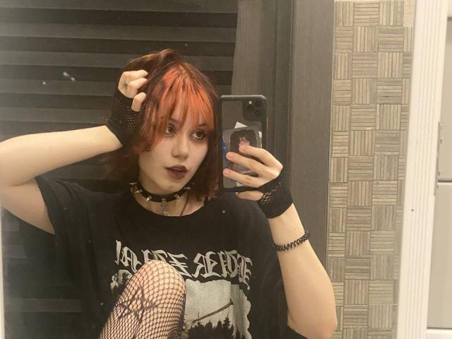 YasukoCella, avec des cheveux courts roux-orangés, un collier noir, du rouge à lèvres foncé, des bas en filet, un t-shirt noir, des gants noirs sans doigts, prenant un selfie devant un miroir, tenant un téléphone avec un support pop..