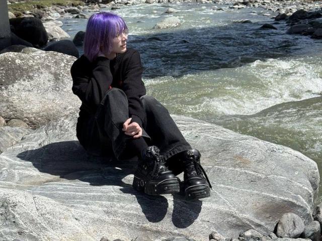 YasukoCella with purple bob sitting on grey rocks, wearing black clothes and boots, hands on knees, looking to the side, by a flowing river with rocks in background.