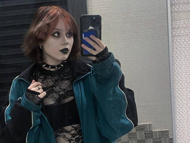 YasukoCella, avec ses courts cheveux roux, son haut en dentelle noire, sa veste turquoise, son collier noir et son rouge à lèvres noir, prend un selfie devant un miroir, tenant un téléphone bleu et montrant ses doigts..