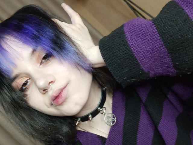 YasukoCella hat lila und schwarzes Haar, einen Nasenring, einen Lippenring und trägt ein schwarz-lila gestreiftes Hemd mit einem Pentagramm-Kragen. Sie berührt ihren Kopf..