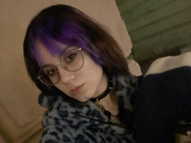 YasukoCella met paars en zwart haar, een bril, een neusring en een lipring, gekleed in een blauwe hoodie met luipaardprint en een zwarte choker, die naar de camera kijkt..