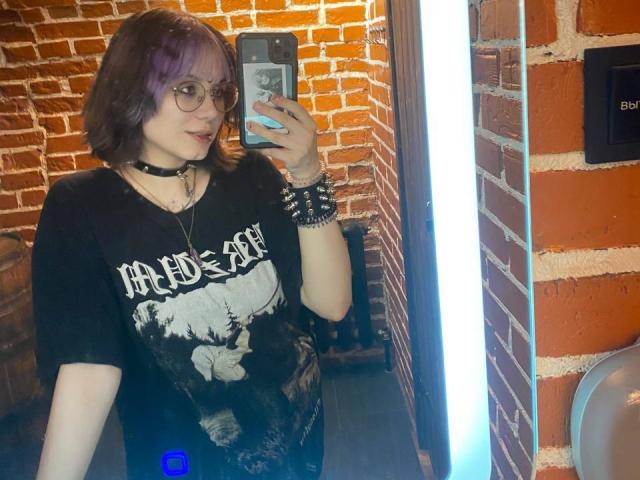 Yasuko, con capelli viola, occhiali, collana nera, maglietta con la scritta "Aiders" e braccialetti con borchie, sta scattando un selfie allo specchio, tenendo un telefono, con uno sfondo di un muro di mattoni e una luce fluorescente a destra..