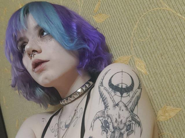 YasukoCella with purple-blue hair, nose ring, freckles, dark lipstick, choker, and tattoos, including a horned skull on her shoulder, looking up. Her tits are partially visible.