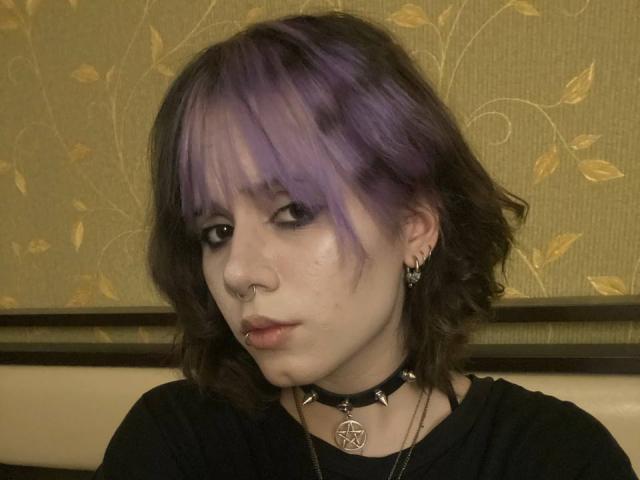 YasukoCella tem cabelo roxo e castanho, delineador preto, piercing no nariz, brincos pequenos, um colar preto com um pentagrama e uma camisa preta. Ela está olhando para baixo com uma expressão levemente emburrada..