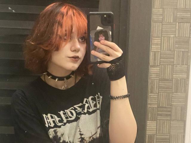 YasukoCella mit roten, gewellten Haaren, einem schwarzen Choker, Gothic-Make-up, einem schwarzen T-Shirt mit weißem Aufdruck, einem schwarzen Handschuh ohne Finger und einem Smartphone, das sie für ein Selfie vor einem Spiegel benutzt. Blasse Haut, ein leichtes Lächeln..