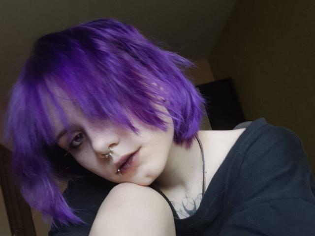 Yasuko, con i capelli viola, un anello al naso, un piercing al labbro, una maglietta nera, pelle chiara e un tatuaggio, guarda la fotocamera con un leggero sorriso. Il suo seno è piccolo e la sua vulva non è visibile..