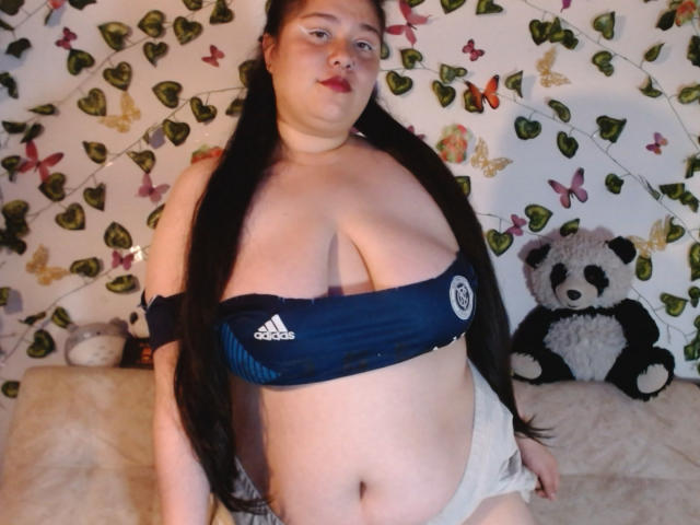 Lena Torner met grote, prominente borsten in een marineblauwe Adidas sportbeha, lange zwarte vlechtjes en beige shorts, staand voor een opgezette panda en een groene wand..