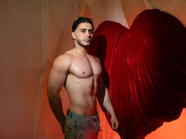 TaylerDuster est un homme musclé, torse nu, avec une poitrine et des abdominaux bien dessinés, portant un jean déchiré, debout à côté d'un tissu rouge drapé, avec une expression sérieuse..