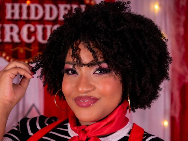 ViaraMoon with curly black hair, red scarf, hoop earrings, pink lipstick, and striped shirt, smiling at the camera, hand near her head. Boobs not visible. Red and white background.