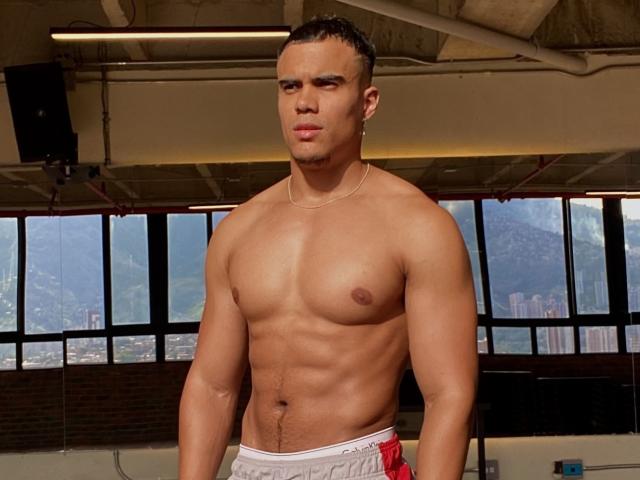 Dominic Salazar est un homme musclé, torse nu, avec une mâchoire forte, des cheveux courts et foncés, et une peau bronzée. Il porte des sous-vêtements blancs et rouges et se tient dans une pièce baignée de soleil..