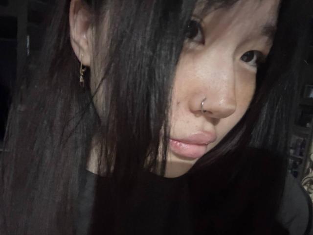 JungMcgranor has long black hair covering one eye, a nose ring, and a small lip ring, wearing a black shirt, with dark earrings, in a dimly lit photo.