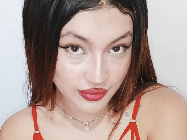 Brenda Ferrer, con lápiz labial rojo, cejas negras y ojos marrones, lleva un sujetador rojo, un collar con un pequeño colgante y cabello negro con puntas marrones. Sus senos son visibles..