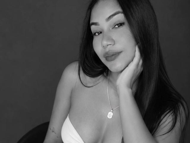 Photo en noir et blanc de liacurve avec de longs cheveux foncés, des lèvres pulpeuses et un collier, reposant sa joue sur sa main, montrant ses seins et un petit tatouage sur son bras..
