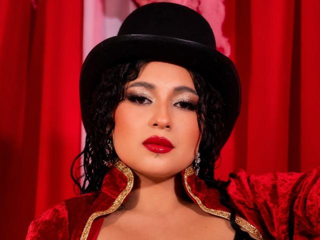 LilithStygian a les cheveux noirs bouclés, porte un chapeau noir, une veste rouge en velours avec des détails dorés, et du rouge à lèvres, avec un maquillage scintillant pour les yeux, et elle a l'air confiante et séduisante..