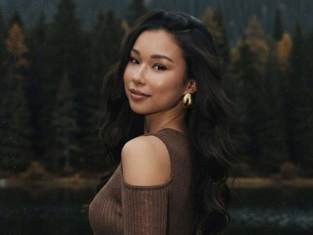 NadineYauger with long wavy dark hair, brown off-shoulder top, gold earrings, smiling at camera, forest in background, looking over shoulder, showing her tits slightly.