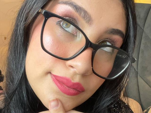 NinaRouse has tan skin, black hair, green eyes, pink eyeshadow, thick black eyebrows, red lipstick, and wears black glasses with a slightly upturned nose, looking cute.