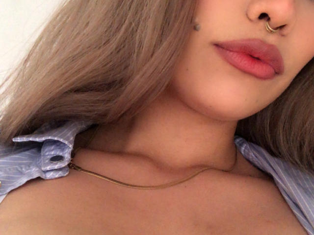 Ilaynia with light brown hair, pinkish-red lips, nose ring, and small black mole, wearing a blue striped shirt and thin gold necklace, showing her upper chest and neck.