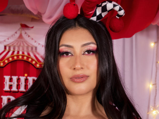 Catalina Rivero, met lang zwart haar, roze oogschaduw, glanzende roze lippen, en een rode en zwarte geruite narrenhoed met pompons, die recht in de camera kijkt..