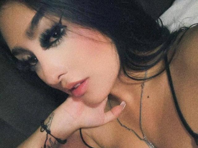 CatalinaNix with dark hair, heavy black eyeliner, and pink lips, her hand touching her neck, showing a tattoo on her finger, wearing a black strap top.