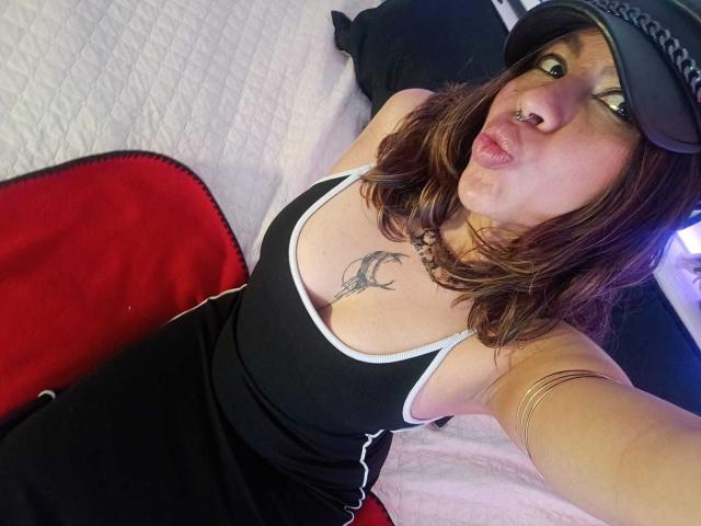 DulceBom with brown hair, wearing a black tank top and black cap, pouting her lips, showing her tits and a tattoo above her cleavage, lying on a bed.