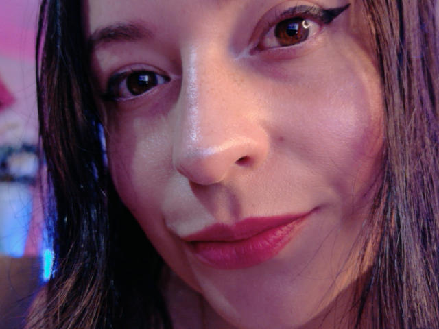 O rosto de VaaleryStark, com olhos castanhos, delineador preto, batom rosado-avermelhado, pele levemente brilhante, nariz pequeno e cabelo castanho, sorrindo gentilmente. Seios não visíveis, foco nas características faciais..