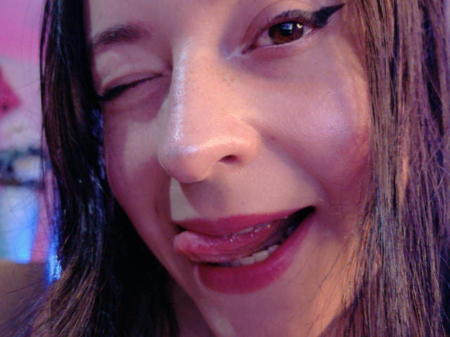 VaaleryStark's face, winking with tongue out, showing red lipstick and black eyeliner, brown eyes, and slightly messy brown hair. Her nose and lips are centered in the shot.