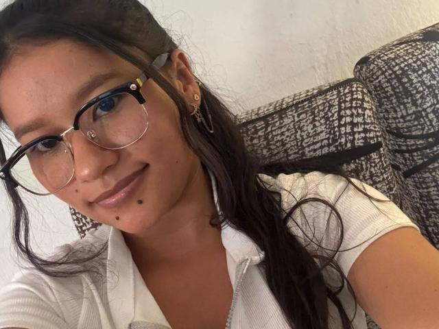 LexieMorgan with glasses, brown hair, white shirt, small mole on chin, smiling, wearing small earrings, sitting on a patterned gray sofa, upper body shot.
