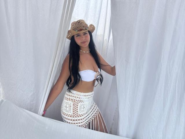 EimyRiverax, avec ses longs cheveux noirs, sa peau bronzée, portant un haut bandeau blanc, une jupe en crochet et un chapeau de paille, se tient devant des rideaux blancs, montrant ses fesses et ses petites seins..