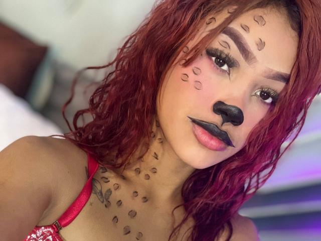 SiennaValois with red wavy hair, leopard print face and nose, black lipstick, red bra, and leopard tattoos on neck, looking at camera, soft lighting, close-up.
